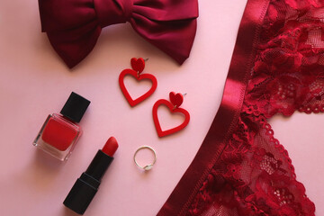 Red lace bralette, hairbow, heart shaped earrings, golden ring, red lipstick and nail polish on pink background. Coquettish accessories for Valentine's day. Flat lay.