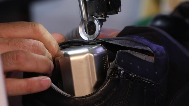 
Close-up view of a shoemaker's hands using an industrial sewing machine to stitch black leather boots in a professional footwear manufacturing factory workshop production line.

