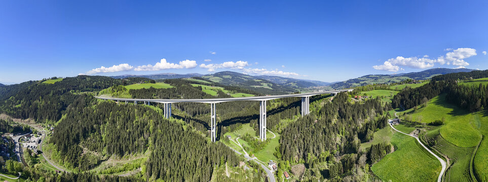 Aerial view of a colossal bridge piercing through a verdant valley, its stark grey contrasting with the lush green hills, Wolfsberg, Carinthia, Austria.