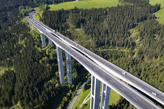 Aerial view of a towering concrete viaduct slicing through a dense forest, where cars traverse the skyway amidst the verdant landscape, Wolfsberg, Carinthia, Austria.