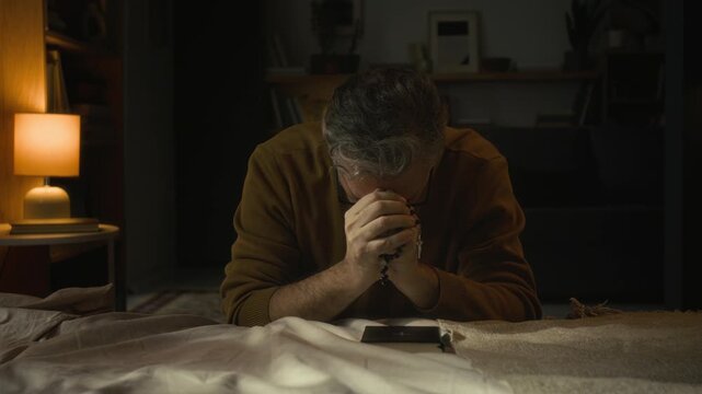 Zoom in view of hopeful Caucasian middle-aged man with clasped hands lowering his head while praying for divine help leaning on edge of bed with elbows