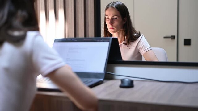 Young businesswoman with a serious expression concentrating on her laptop screen while sitting at her home office desk. Her face reflects in a portable monitor as she works on a project