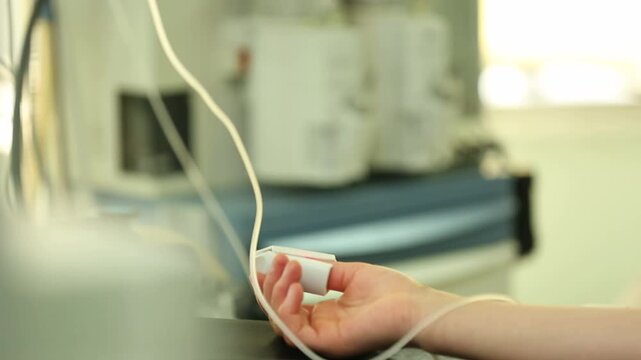 A patient lies in an operating room with a pulse oximeter on their finger and an IV line in their arm while medical staff prepare equipment nearby.

