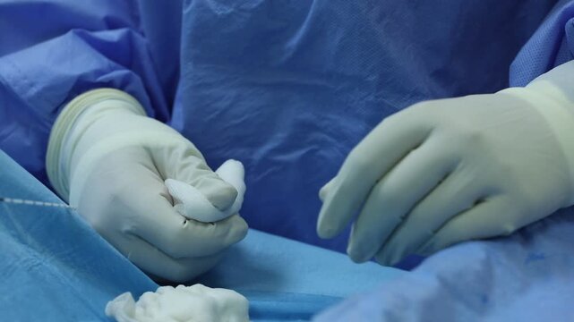 Close-up of professional surgeon hands in sterile gloves holding gauze and guiding a thin medical wire instrument during a precise surgical procedure in a hospital operating room environment.

