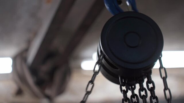 Close-up of a black chain hoist pulley system slowly moving in an industrial warehouse. The focus is on the mechanism lifting heavy equipment in a factory or construction site