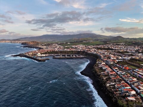 Aerial view of a rugged coastline where the dark volcanic sand meets the turbulent ocean, and buildings cluster along the shore beneath rolling hills, Rabo de Peixe, Azores, Portugal.