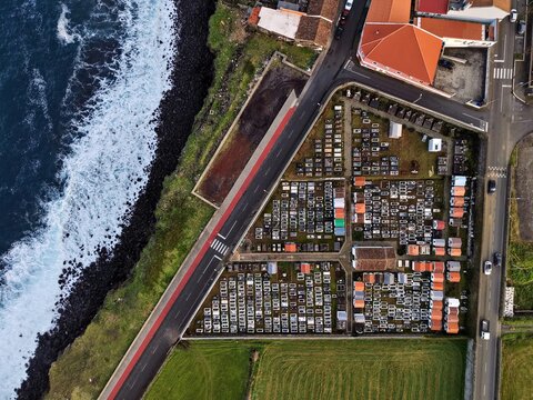 Aerial view of a cemetery with rows of graves standing in solemn silence, juxtaposed against the restless ocean waves crashing on the dark volcanic coastline, Rabo de Peixe, Azores, Portugal.