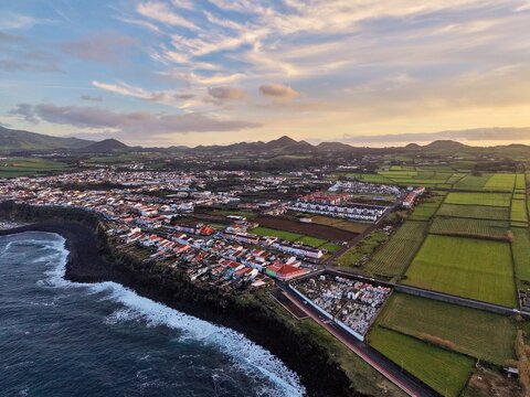 Aerial view of the coastal town where the dark volcanic rocks meet the azure ocean, contrasting with the green fields and distant mountains, Rabo de Peixe, Azores, Portugal.