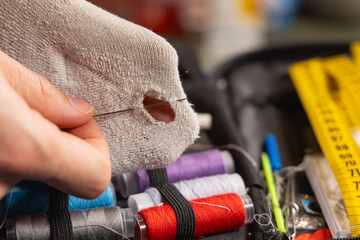 Une personne en train de réparer un trou dans une chaussette avec une aiguille inséré et du fil. En arrière plan, une trousse à couture avec des bobines de fil. © Gautier Normand