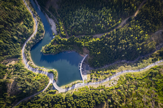Aerial view of a dam nestled between a winding river and dense green forests, casting shadows over the water's surface, Bodendorf, Styria, Austria.