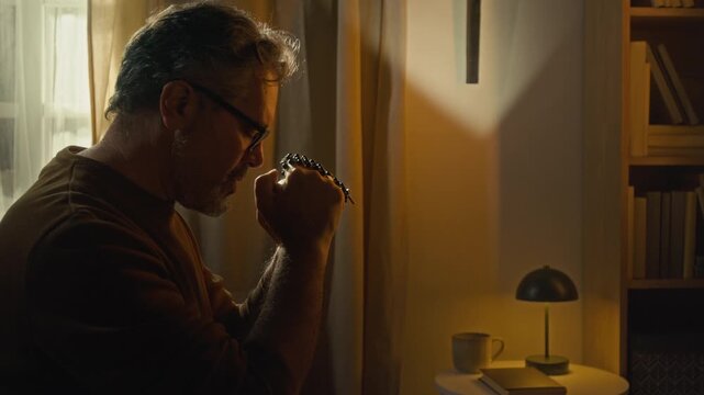 Tilt up side view of Caucasian mature man raising joined hands to his face while praying to God, bedside table with mug, book and lamp along with cross on wall in background