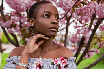 Beautiful young black woman with short hair, wearing a floral dress, is posing with her hands on her face and head, under a blooming cherry blossom tree in spring. Japanese Sakura Flower