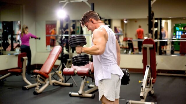 Young man is focused on lifting weights in a gym filled with people using workout equipment during daytime hours