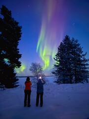 Two girls standing in front of Northern Lights and taking pictures of this majestic natural phenomenon during ice cold polar night close to the cities of Inari and Ivalo in  Lapland, Finland © been.there.recently