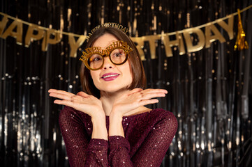 Fototapeta premium Cute and elegant portrait of an adult woman in a red glitter dress wearing novelty glasses with “Happy Birthday” text on dark festive background. Concept of joyful celebration, charm, femininity