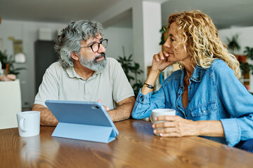 Portrait of a lovely tablet senior mature couple using a laptop together and having fun drinking coffee or tea sitting at a table at home