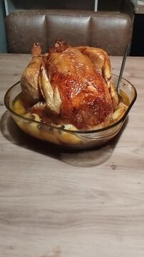 Roasted capon resting in a transparent glass dish on a wooden table, with a metal spoon left inside the dish, bathing in cooking juices, traditional homemade cooking scene.