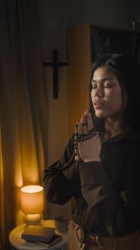 Vertical rack focus view of devoted Caucasian gen Z girl dressed in shirt and pants turning to God in prayer while standing with her hands folded together in dimly lit room with cross on wall
