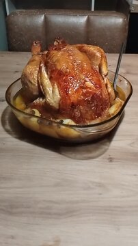 Roasted capon resting in a transparent glass dish on a wooden table, with a metal spoon left inside the dish, bathing in cooking juices, traditional homemade cooking scene.