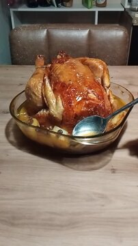 Roasted capon resting in a transparent glass dish on a wooden table, being basted with its cooking juices using a metal spoon, traditional homemade cooking scene.