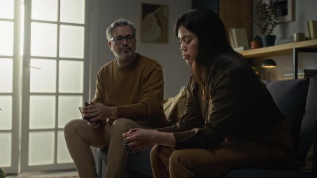Devastated young woman with rosary looking down and sharing her feelings with mature spiritual guide who holding bible book and carefully listening