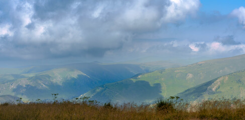 Fototapeta premium Wide view of green hills and mountains under cloudy blue sky.Expansive landscape with layered green hills and distant mountains under a cloudy blue sky. Wild plants in foreground