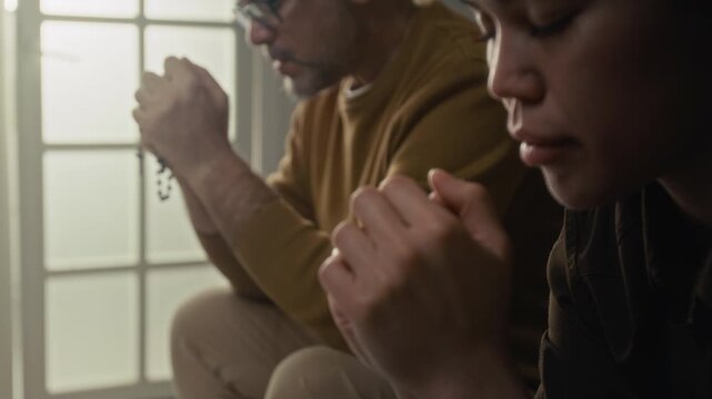 Close-up rack focus view of young woman whispering prayer with eyes closed while praying next to mature spiritual mentor holding rosary