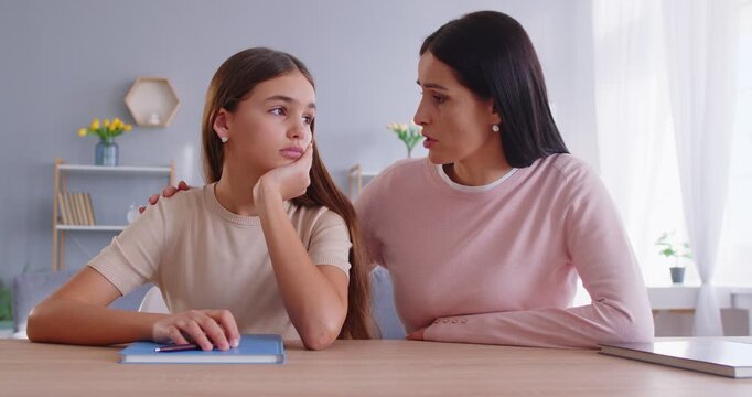 Caring worried mother comforting her upset teenage daughter sitting at table. Concerned parent offering support and empathy to her child who telling about her problems and difficult life situation.