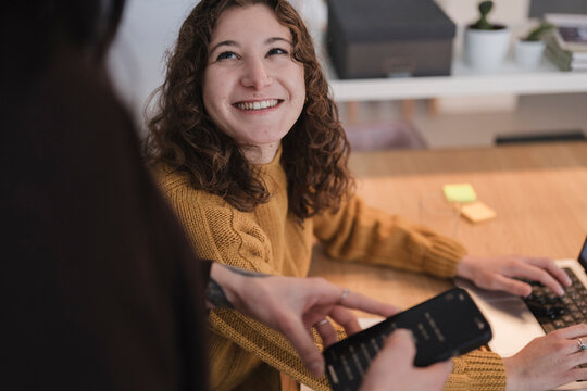 A young woman with curly hair smiles while working on a laptop and holding a remote control.