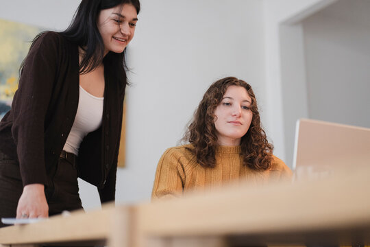 Two young women collaborate on a project, one looking at a laptop while the other leans in to observe and offer input.