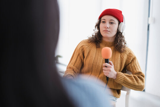A young woman wearing headphones and a red hat holds a microphone, engaging in an interview or recording session.