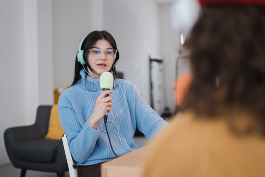 A young woman wearing headphones and glasses speaks into a microphone while recording a podcast with another person.