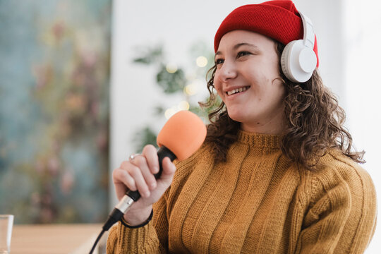 A young woman wearing headphones and a red beanie smiles while holding a microphone, ready to record her podcast or broadcast.