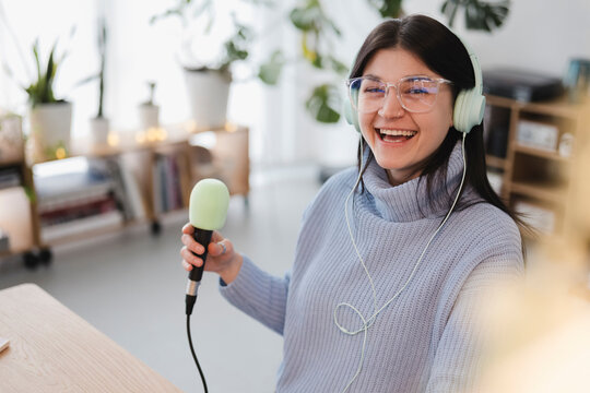 A young woman wearing headphones and glasses laughs while holding a microphone, ready to record her podcast or broadcast.