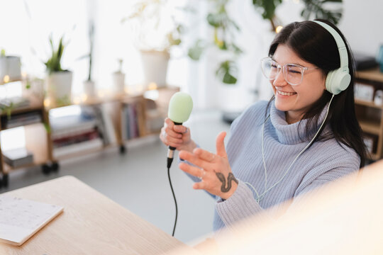 A young woman with headphones and glasses smiles as she holds a microphone, recording audio in a bright, plant-filled room.