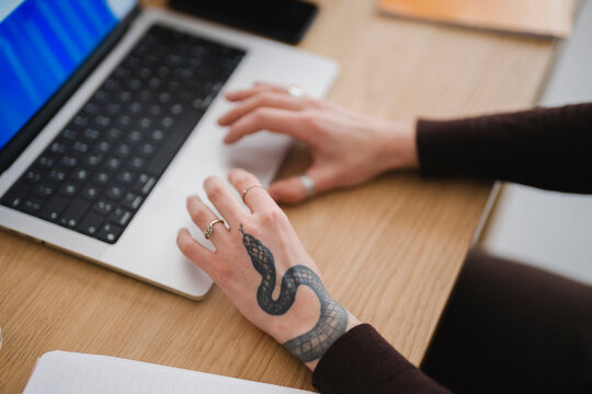 A person with a snake tattoo on their hand types on a laptop, with a notebook open nearby on a wooden desk.