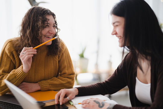 Two young women are studying together, laughing and interacting while working on their assignments at a table.
