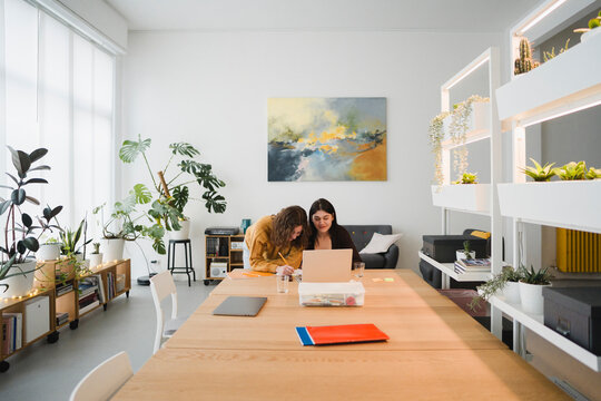 Two young women collaborate on a project at a wooden table in a bright, modern office space filled with plants.