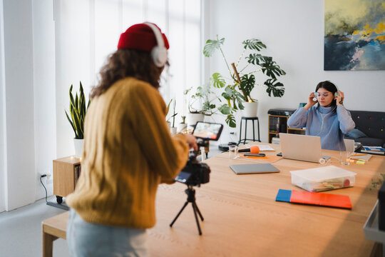 Two women are in a bright room, one recording with a camera and the other working on a laptop, both wearing headphones.