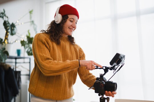 A young woman wearing headphones and a red beanie adjusts a professional camera on a tripod, preparing to film content.