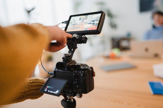 A person's hand adjusts a camera monitor, capturing a scene with a person working on a laptop in the background.