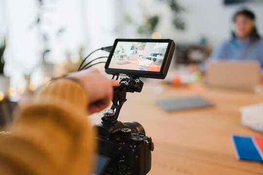 A person's hand adjusts a camera monitor, capturing a video call with another person working on a laptop in the background.