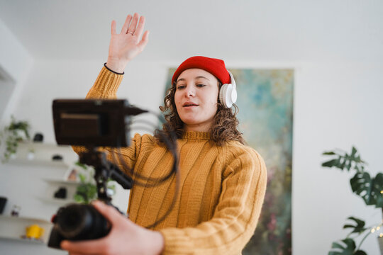 A young woman wearing headphones and a red beanie records a video with a camera on a tripod, waving hello to her audience.