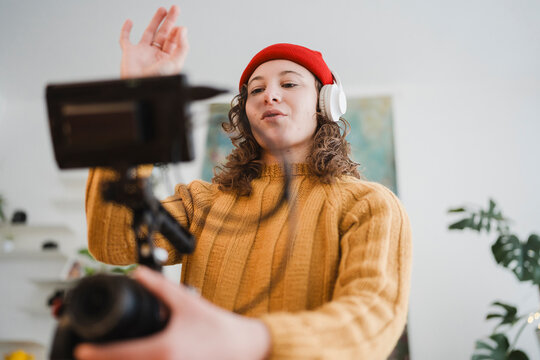 A young woman wearing a red hat and headphones records a video on a camera set up on a tripod, gesturing with her hand.
