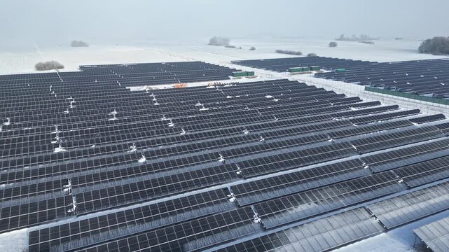 Aerial view of expansive solar panel farm covered in snow, showcasing rows of solar panels and distant fields under a cloudy sky in winter landscape