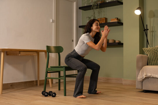 Smiling athletic woman performing chair squat exercise at home using bodyweight training to strengthen legs glutes and core during indoor functional fitness workout