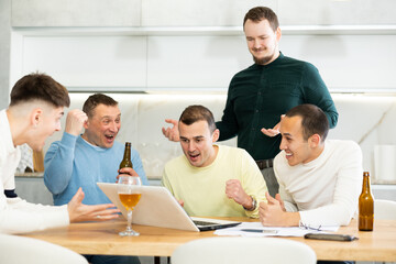 Fototapeta premium Group of men watching sports match online on laptop during friendly meeting with beer at home, excitedly celebrating winning score in game ..