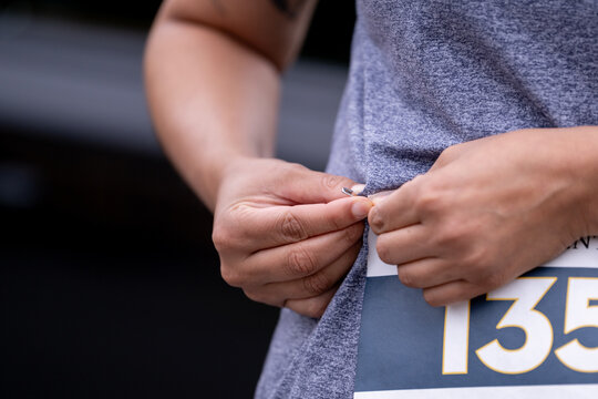 Rrunner's hands carefully attaching race bib number to t-shirt using safety pin in preparation for race or marathon event. Shows pre-race ritual and participant preparation.  - Powered by Adobe