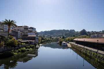 Fototapeta premium Scenic view of Ponte Vella bridge over Mandeo River in Betanzos, Galicia, Spain, surrounded by lush greenery and historic architecture.