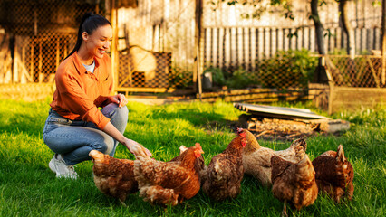 Young woman feeding her chickens kneeling on the grass, hens walking on farm backyard, side view shot © Home-stock